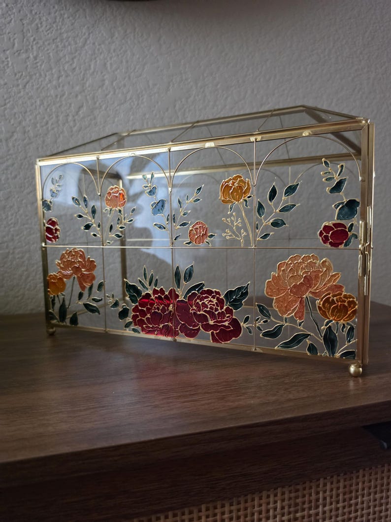 May include: A rectangular glass display box with a gold metal frame. The glass panels feature hand-painted floral designs in shades of deep red, orange, and yellow, accented with dark green leaves and thin gold outlines throughout the structure.