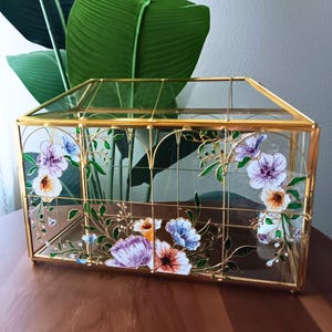 May include: A rectangular glass display box with a gold metal frame. The glass panels are decorated with hand-painted floral motifs in shades of purple, blue, orange, and white, featuring green leaves and thin vines that follow the metal grid lines.