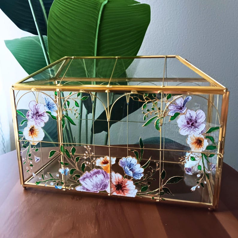May include: A rectangular glass display box with a gold metal frame. The glass panels are decorated with hand-painted floral motifs in shades of purple, blue, orange, and white, featuring green leaves and thin vines that follow the metal grid lines.