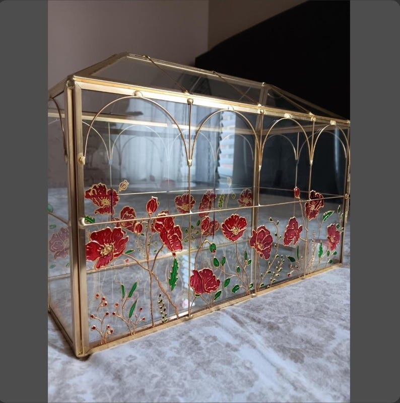 May include: A rectangular glass display box with a gold metal frame and a gabled roof. The clear glass panels are decorated with hand-painted red poppy flowers, green stems, and small buds. The structure features arched metal detailing along the top.