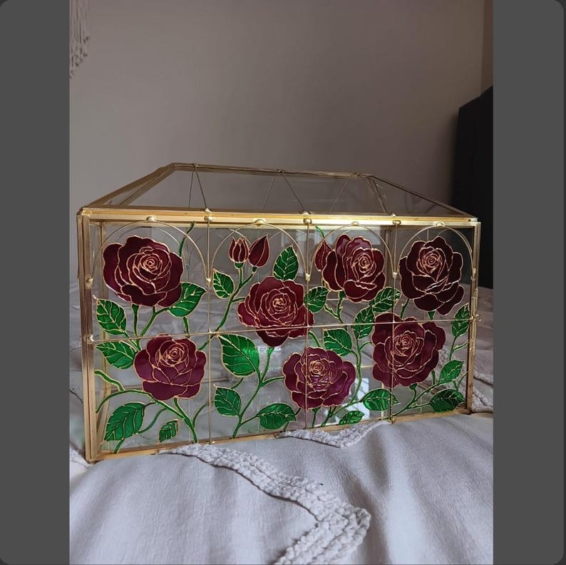 May include: A rectangular glass display box with a gold metal frame. The front glass panel features a hand-painted design of deep red roses with green leaves and stems, outlined in gold. The box has a hinged lid and clear glass sides.