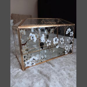 May include: A rectangular glass display box with a gold metal frame, featuring hand-painted white and black floral branch designs on the front panels. The top glass panel is inscribed with the name Monique in elegant cursive gold lettering.