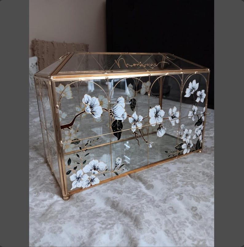 May include: A rectangular glass display box with a gold metal frame, featuring hand-painted white and black floral branch designs on the front panels. The top glass panel is inscribed with the name Monique in elegant cursive gold lettering.