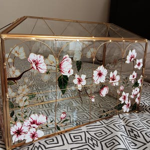 May include: A rectangular glass display box with a gold metal frame. The glass panels are decorated with hand-painted white and pink cherry blossom flowers on brown branches with green leaves. The structure features a hinged lid and clear glass sides.
