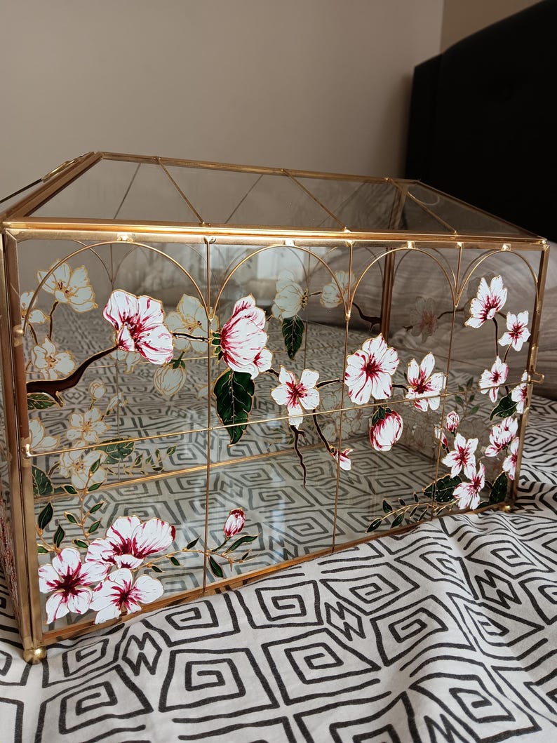 May include: A rectangular glass display box with a gold metal frame. The glass panels are decorated with hand-painted white and pink cherry blossom flowers on brown branches with green leaves. The structure features a hinged lid and clear glass sides.