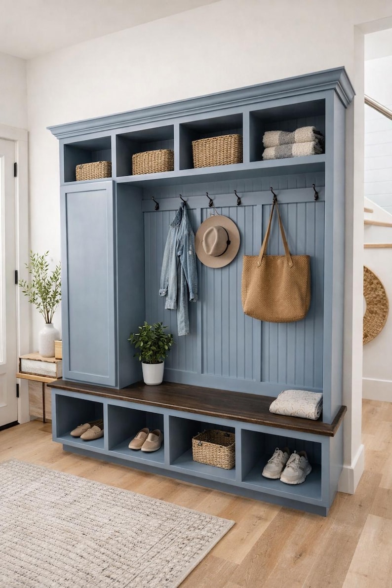 May include: A blue entryway storage unit with a bench, shoe cubbies, and upper shelves. Wicker baskets, a denim jacket, a hat, and a tote bag hang from hooks. A rug and shoes are in the foreground.