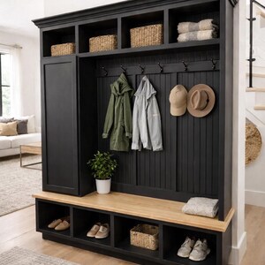 May include: A black entryway storage unit with a bench, shoe cubbies, and upper shelves. Wicker baskets, coats, and hats hang from hooks. A small potted plant and folded blanket add decor. The unit is made of wood.