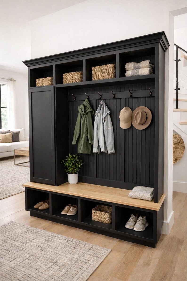 May include: A black entryway storage unit with a bench, shoe cubbies, and upper shelves. Wicker baskets, coats, and hats hang from hooks. A small potted plant and folded blanket add decor. The unit is made of wood.