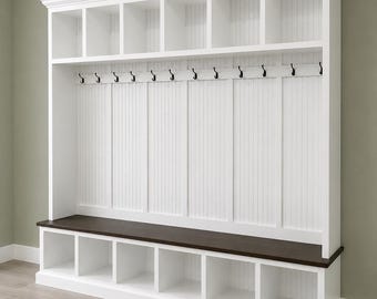 May include: A white wooden entryway storage unit with multiple cubbies, a bench, and coat hooks. The unit has a dark brown bench top and a series of open storage compartments above and below the bench. The wall behind the hooks has vertical paneling.
