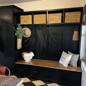 Black entryway storage bench with three drawers, cubbies, and hooks.  Features a built-in cabinet and woven baskets. The bench has a light brown top and includes a pillow with the text 'Athens GA'.  The overall style is farmhouse or rustic.