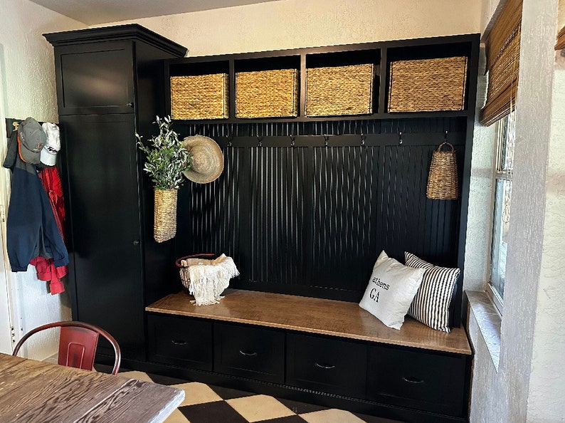 Black entryway storage bench with three drawers, cubbies, and hooks.  Features a built-in cabinet and woven baskets. The bench has a light brown top and includes a pillow with the text 'Athens GA'.  The overall style is farmhouse or rustic.