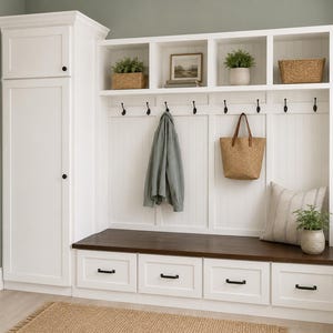 May include: A white entryway storage unit with a dark brown bench and four drawers. Above, there are open shelves with decorative items, hooks for hanging coats, and a woven tote bag. A framed botanical print hangs on the wall.