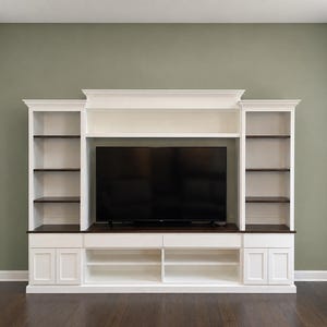 May include: A white entertainment center with a large black television in the center. The unit has built-in bookshelves on either side with dark brown shelves. The bottom section has cabinets and open shelving.