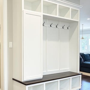 May include: A white built-in entryway storage unit with a dark brown bench seat. The unit features open cubbies, a cabinet, and hooks for hanging items. The design includes crown molding and is set against a light gray wall.