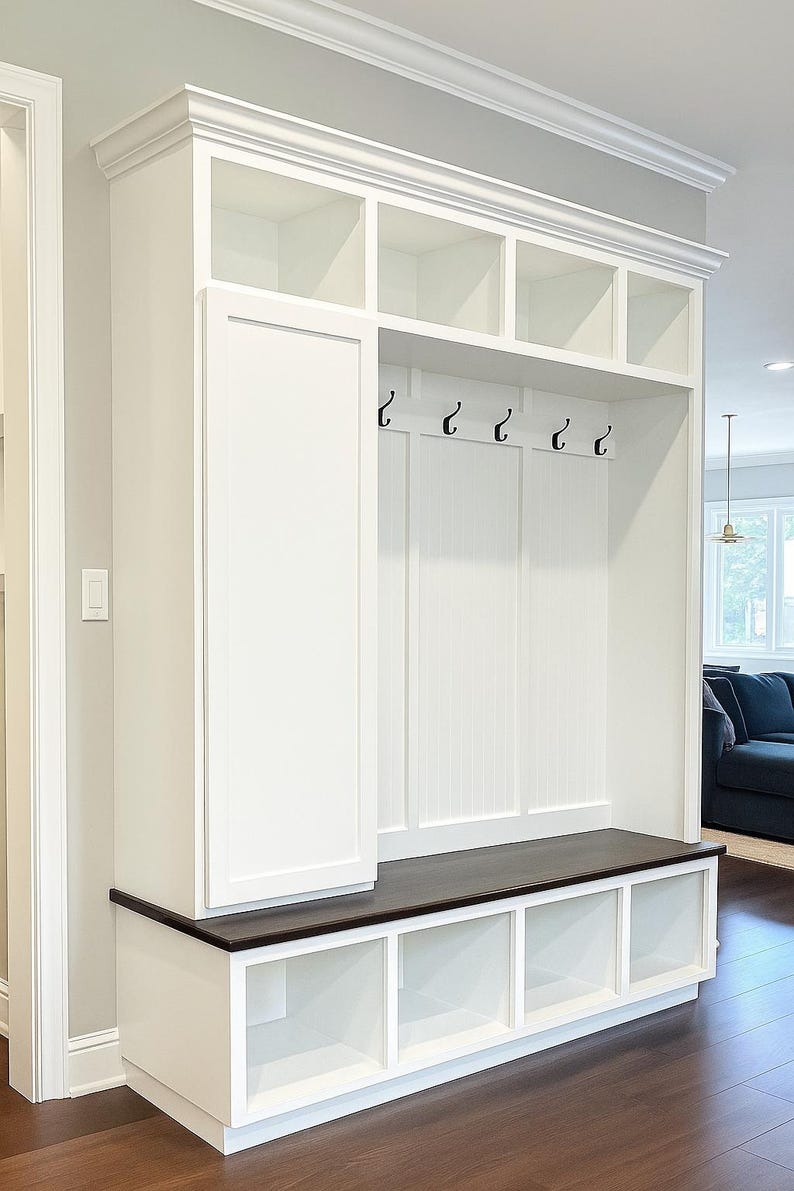 May include: A white built-in entryway storage unit with a dark brown bench seat. The unit features open cubbies, a cabinet, and hooks for hanging items. The design includes crown molding and is set against a light gray wall.