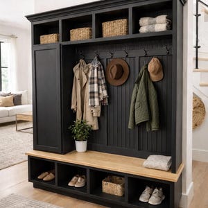 May include: A black entryway storage unit with a bench, shoe cubbies, and upper shelves. Woven baskets and folded blankets are stored on the shelves. Coats, hats, and a scarf hang from hooks. A small potted plant sits on the bench.