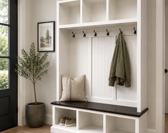 May include: A white entryway storage unit with a dark wood bench and open cubbies below. The unit has a series of hooks for hanging coats, a decorative pillow, and a framed picture. A green jacket hangs on one of the hooks.