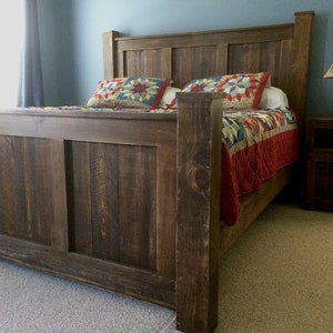 May include: A rustic wooden bed frame with a dark brown finish. The headboard has a simple design with vertical planks. The bed is made up with a red and blue quilt.
