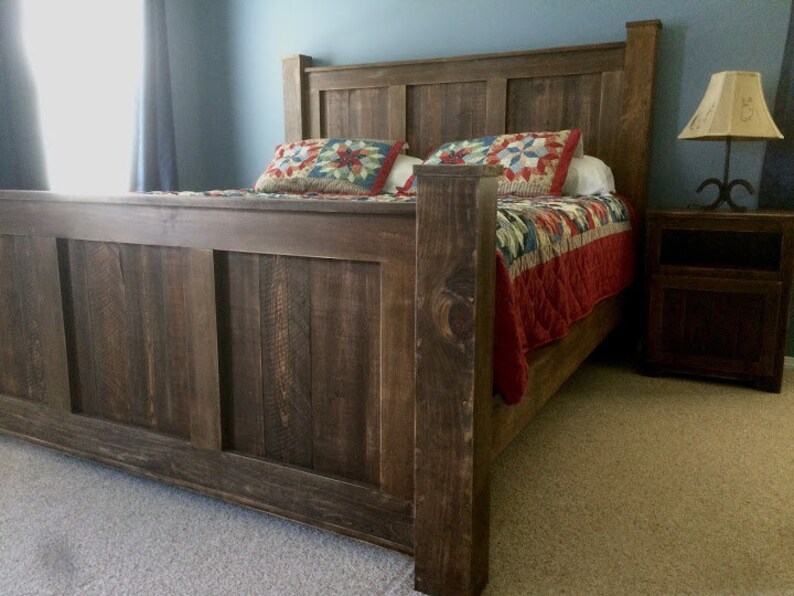 May include: A rustic wooden bed frame with a dark brown finish. The headboard has a simple design with vertical planks. The bed is made up with a red and blue quilt.