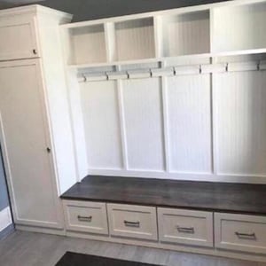 White built-in storage unit with a bench seat and drawers. The unit features cubbies, hooks, and a dark brown countertop.  The cabinet is made of white wood with a shaker style. The unit is ideal for entryways or mudrooms, offering ample storage and organization.