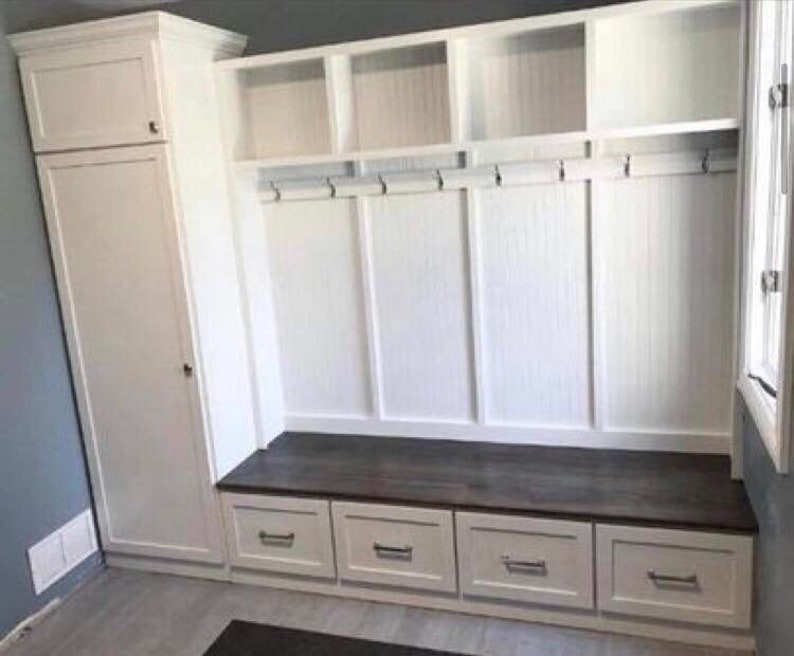White built-in storage unit with a bench seat and drawers. The unit features cubbies, hooks, and a dark brown countertop.  The cabinet is made of white wood with a shaker style. The unit is ideal for entryways or mudrooms, offering ample storage and organization.