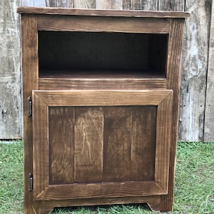 May include: A dark brown wooden cabinet with a single door and a shelf. The cabinet has a simple design with a slightly distressed finish.