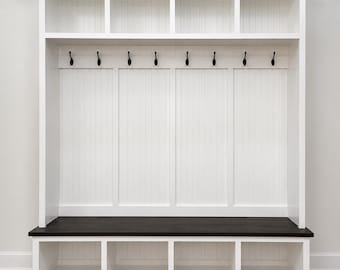 May include: A white wooden hallway storage unit with a dark brown bench seat. It has open cubbies at the top, six black hooks, and lower storage compartments. The unit is designed for storage and has a clean, modern aesthetic.