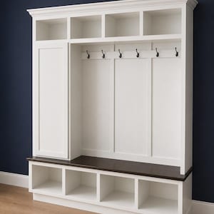 May include: A white wooden entryway organizer with a dark brown bench seat. It features open storage cubbies, a cabinet, and hooks for hanging items. The unit is against a dark blue wall.