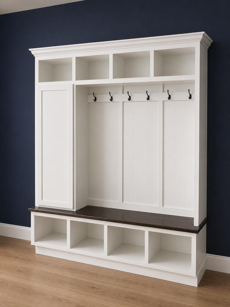 May include: A white wooden entryway organizer with a dark brown bench seat. It features open storage cubbies, a cabinet, and hooks for hanging items. The unit is against a dark blue wall.
