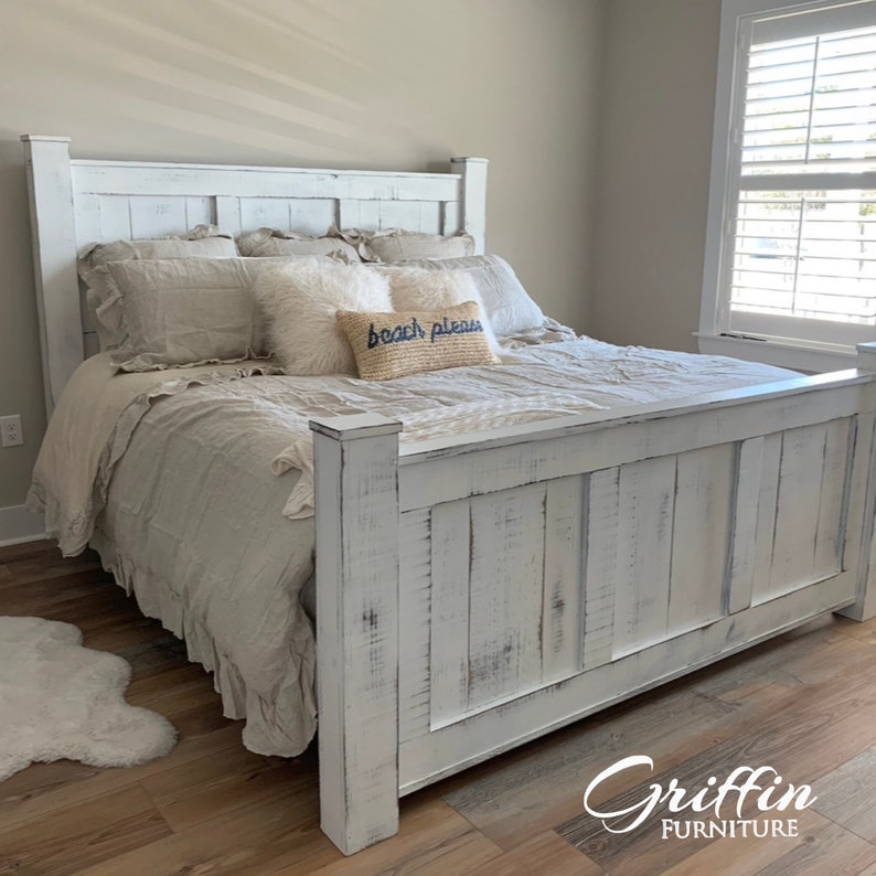 May include: White wooden bed frame with a distressed finish. The headboard has a simple design with vertical planks. The bed is made up with a white and beige bedding set and a pillow with the text "beach please".