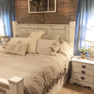 May include: A white bed frame with a beige linen duvet and ruffled trim. The bed is decorated with pillows, including one with the text "I LOVE US". The headboard is against a wood wall with a window above it.