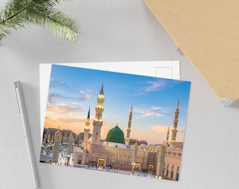 May include: A rectangular postcard featuring a high-angle photograph of the Al-Masjid an-Nabawi mosque with its iconic green dome and tall minarets illuminated against a soft sunset sky, positioned next to a silver pen and a pine branch.