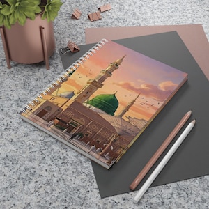 Pode incluir: Caderno espiral com uma ilustração detalhada de uma mesquita com uma cúpula verde e minaretes sob um céu de pôr do sol. O caderno está sobre uma superfície cinza com lápis e uma pequena planta no fundo.