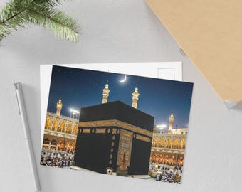 May include: A postcard featuring the Kaaba in Mecca, Saudi Arabia, under a starry night sky with a crescent moon. The black cube-shaped structure is adorned with gold accents, surrounded by a crowd of people and illuminated buildings.
