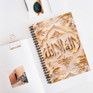 May include: A spiral-bound notebook featuring a cover with gold Arabic calligraphy set against a cream-colored background with gold floral accents and marble textures. The notebook is open, revealing a white page with a small square photograph.