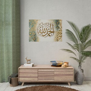 May include: A framed artwork featuring Arabic calligraphy in gold against a white background with teal and gold decorative patterns. The artwork hangs above a wooden cabinet with a decorative bowl, candles, and a plant.