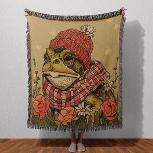 May include: A woven throw blanket featuring a green toad wearing a red knit beanie with a white flower and a red and white plaid scarf. The background is a tan color with illustrations of orange flowers and white daisies with green stems.