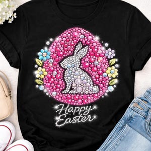 Könnte beinhalten: Schwarzes T-Shirt mit einem Oster-Design. Das Design zeigt einen weißen Hasen in einem rosa Ei, umgeben von bunten Blumen. Darunter steht in Glitzer-Schrift "Happy Easter".