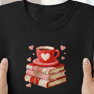 May include: Black t-shirt featuring a stack of books with a red teacup on top. The teacup has a white heart design. Pink hearts are scattered around the books and teacup. The books have a quilted pattern and are tied with a bow.