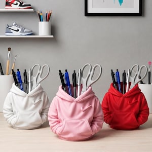 May include: Three custom 3D printed pen holders shaped like miniature hoodies in white, pink, and red. Each hoodie features a small swoosh logo on the chest and a front kangaroo pocket, filled with various pens, pencils, and a pair of scissors.