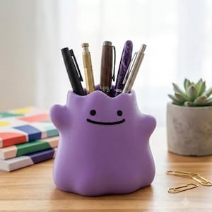 May include: A purple, blob-shaped desk organizer featuring a simple face with two black dot eyes and a thin, curved black mouth. The container is filled with five various pens and pencils, including black, metallic, wooden, and purple patterned designs.