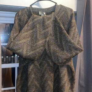 May include: A brown and green patterned sweater with a square neckline and short sleeves hangs on a black hanger. The sweater has a textured, woven appearance and a chevron design. The garment is displayed in front of a window.