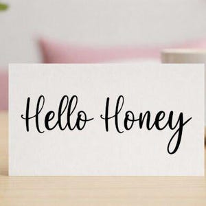 Può includere: Un cartoncino rettangolare bianco con il testo Hello Honey scritto in un carattere corsivo nero. Il cartoncino è posizionato orizzontalmente e presenta un design pulito e semplice con la scritta centrata sulla superficie bianca.