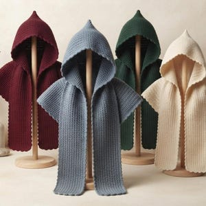 Könnte beinhalten: Vier Kapuzenumhänge in Burgunderrot, Schieferblau, Waldgrün und Creme, jeweils mit einem strukturierten Strickmuster und gewellten Kanten. Diese Kleidungsstücke werden auf Holzständern präsentiert und zeigen eine spitze Kapuze.