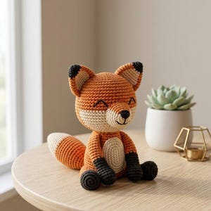 May include: A crochet fox toy featuring orange, cream, and dark brown yarn. The fox has pointed ears with dark tips, a cream-coloured muzzle and belly, and dark brown paws. It is depicted in a seated position with a bushy tail extending to the side.