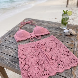 Puede incluir: Un conjunto de bikini de ganchillo en color rosa empolvado que consta de un top triangular con tiras al cuello y una falda a juego con un patrón de motivos circulares y cintura con cordón. El conjunto está hecho de hilo con un dobladillo festoneado.