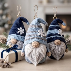 Könnte beinhalten: Drei gehäkelte Wichtel-Ornamente mit spitzen Mützen in Marine- und Hellblau, jeweils mit einer weißen Schneeflocken-Applikation, einer runden beigen Nase und einem langen weißen Garnbart. Jeder Wichtel hat einen goldenen Ring zum Aufhängen.