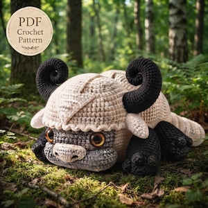 May include: A crochet pattern PDF featuring a beige and grey creature with large black curled horns and brown eyes. The creature has a textured, stitched appearance with a rounded head, a snout, and dark black paws. The text PDF Crochet Pattern is visible.