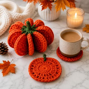 May include: A crocheted orange pumpkin with a green stem, a matching flat circular orange pumpkin coaster, and a speckled ceramic mug filled with a light brown beverage resting on a second orange pumpkin coaster, all on a white marble surface.
