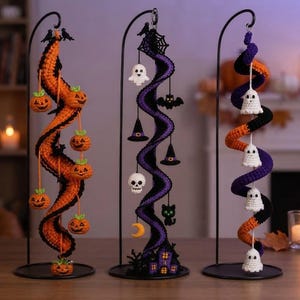 Puede incluir: Tres decoraciones de Halloween de ganchillo en espiral colgadas de soportes de metal negro. La izquierda tiene calabazas naranjas, el centro muestra telaraña, fantasma, murciélago, sombreros, calavera, gato y luna, y la derecha fantasmas blancos.